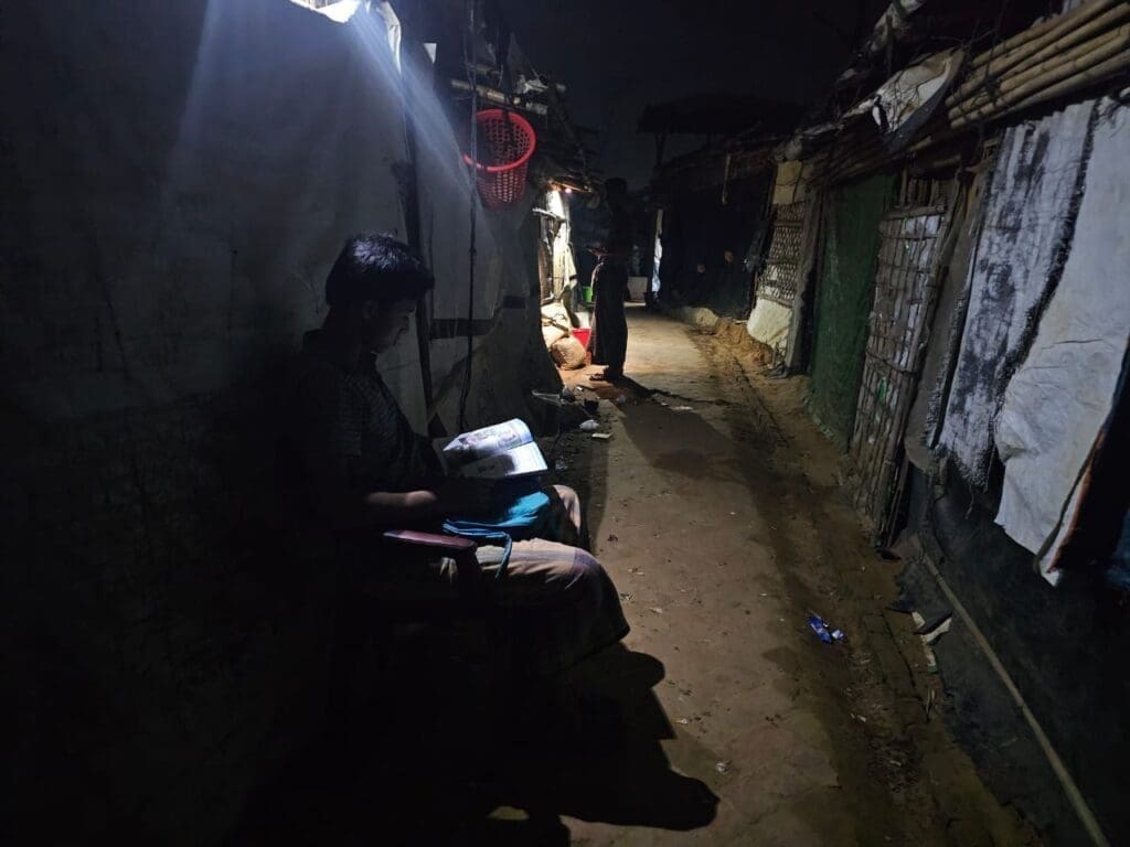 صورة لصبي من الروهينجا يدرس على جانب الطريق تحت ضوء أحد الجيران في مخيم اللاجئين في كوكس بازار على الرغم من الظلام من حوله.